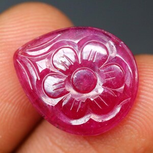May include: A carved ruby gemstone with a floral design. The gemstone is a deep red color and features a detailed carving of a flower with multiple petals. The carving is white, creating contrast against the red stone.