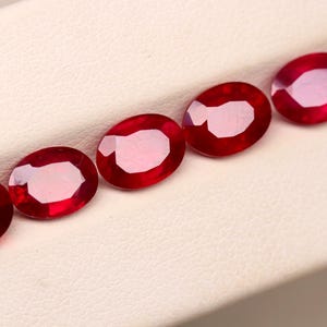 Puede incluir: Cinco piedras preciosas rojas, de corte ovalado, están dispuestas en diagonal sobre una superficie blanca. Las gemas tienen una superficie pulida y reflectante, mostrando su color vibrante y estructura interna. Probablemente son rubíes.