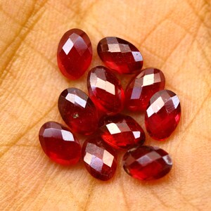 Puede incluir: Un grupo de gemas facetadas, de forma ovalada y de color rojo intenso. Las gemas están dispersas sobre una superficie de color naranja claro, con la luz reflejándose en sus superficies talladas. Las gemas parecen pulidas.
