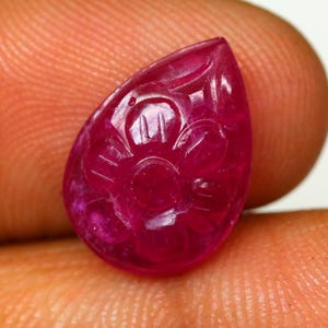 May include: A teardrop-shaped, carved ruby gemstone. The deep red stone features a floral design carved into its surface. The gemstone is held between two fingers, showcasing its size and detail.