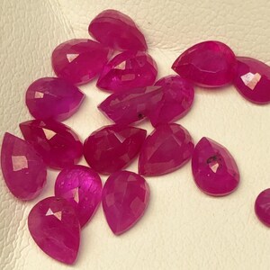May include: A collection of faceted, teardrop-shaped, deep pink gemstones. The gems are scattered on a white surface, each displaying multiple facets that catch the light. The overall effect is a display of vibrant, polished stones.