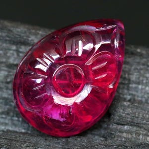 Puede incluir: Una gema tallada en forma de lágrima, de color magenta vibrante. La gema presenta un diseño floral tallado en su superficie, con un elemento central circular y detalles en forma de pétalos. Sobre un fondo oscuro y texturizado.