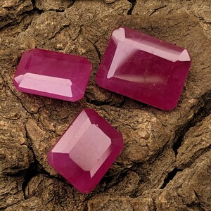 May include: Three faceted, rectangular-cut rubies in varying sizes. The gemstones are a deep, vibrant pinkish-red color, with a polished, reflective surface. They rest on a textured, brown bark background.