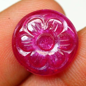 May include: A round, polished, ruby-colored gemstone with a carved floral design. The flower has multiple petals and a central circular element. The gemstone is held between fingers, showcasing its vibrant color and intricate details.