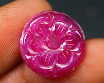 May include: A circular, polished ruby gemstone with a carved floral design. The gemstone is a vibrant pinkish-red colour, with intricate details in the carved flower pattern. The gemstone is held between fingers.