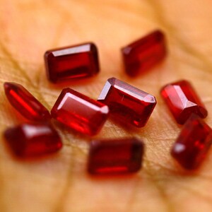 May include: A collection of eight small, deep red gemstones, each cut into a rectangular emerald shape with faceted surfaces that reflect light. The stones are arranged in a cluster, displaying a vibrant, saturated crimson hue throughout.