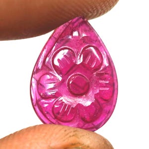 Op de afbeelding: Een gesneden, druppelvormige edelsteen in een levendige magenta tint. De edelsteen heeft een ingewikkeld bloemmotief dat in het oppervlak is gesneden. De edelsteen wordt tussen vingers gehouden tegen een witte achtergrond.