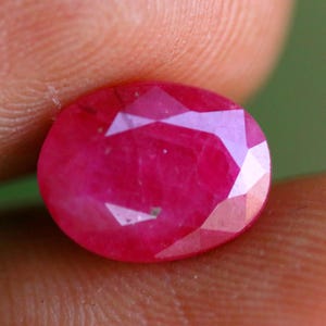 Peut inclure: Une pierre précieuse rubis de taille ovale avec une surface à facettes qui reflète la lumière. La pierre présente une couleur rouge profond et saturée avec des inclusions internes visibles et une finition polie, mettant en valeur sa structure cristalline naturelle.