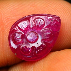 May include: A close-up of a carved, teardrop-shaped ruby gemstone. The gemstone is a deep, vibrant red with intricate floral carvings. The carvings are visible on the surface of the gemstone, adding texture and detail to the design.