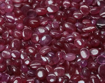 May include: A close-up of a collection of polished, oval gemstones. The stones are a deep burgundy hue with a glossy, reflective surface. The gemstones vary in size and are closely packed together, creating a textured appearance.
