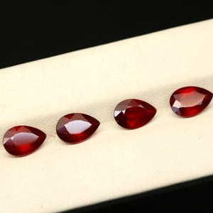 May include: Six faceted, pear-shaped, deep red gemstones are displayed in a row. The gems are arranged on a light beige surface, with a dark background. The gems have a high luster and reflect light, showcasing their cut and clarity.