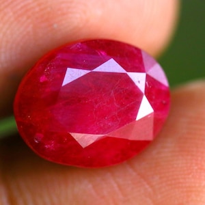 May include: A vibrant, oval-cut ruby gemstone featuring a deep red color and a faceted surface that reflects light. The stone displays internal clarity characteristics and a polished finish, showcasing a classic gemstone shape with symmetrical facets.