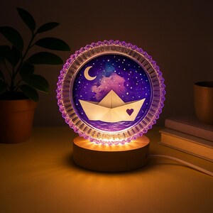 Op de afbeelding: Decoratieve nachtlamp met een papieren bootontwerp in een cirkelvormige lijst. De lijst heeft een paarse kralenrand en staat op een houten voet. De afbeelding toont een nachthemel met een maan en sterren.