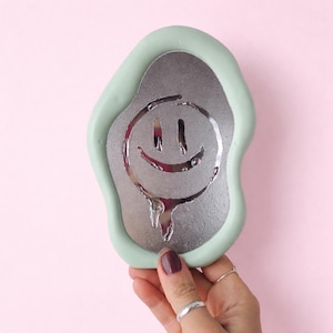 Può includere: Specchio verde menta e argento con un design a faccina sorridente che si scioglie. Lo specchio ha una forma irregolare e organica. La faccina sorridente argentata presenta un effetto gocciolante, su sfondo rosa.