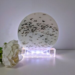 May include: A white circular resin sculpture with a flock of black birds in flight. The sculpture is illuminated from below by a white LED light base.