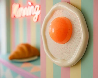 May include: A ceramic fried egg wall art piece with a bright orange yolk and a speckled white egg white. The background features a neon sign that says "Morning" and a croissant.