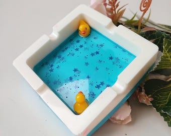 May include: A white ceramic ashtray shaped like a pool with blue water and white stars. Two yellow rubber duckies are floating in the water.