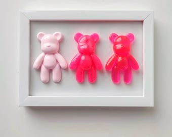 Puede incluir: Tres figuritas de osito de peluche de resina en un marco blanco. El osito de peluche de la izquierda es rosa, el osito de peluche del medio es rosa con purpurina y el osito de peluche de la derecha es rosa con purpurina.