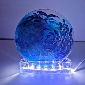 May include: A decorative, illuminated art piece featuring a blue and white swirling design within a clear, rounded form. The piece rests on a rectangular base with embedded lights, creating a soft glow. A white cord extends from the base.
