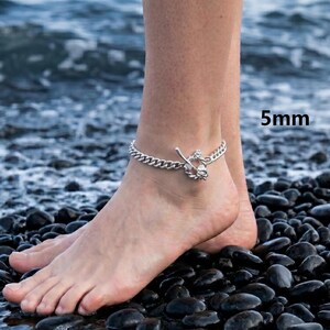 May include: A silver-colored anklet with a chain design and a decorative charm. The anklet is 5mm wide and is worn on the ankle. The background shows a beach with dark rocks and water.