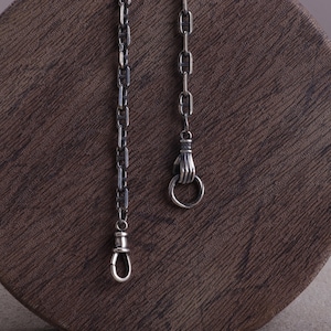 Puede incluir: Primer plano de dos cadenas plateadas. Una cadena tiene un cierre de mosquetón y la otra una mano que sostiene un anillo. Las cadenas se muestran sobre una superficie marrón texturizada. Las cadenas son probablemente para joyería o accesorios.