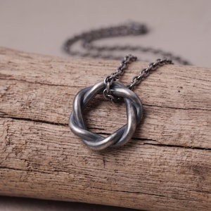 Puede incluir: Un colgante de metal en tono plateado oscuro con forma de nudo circular trenzado, suspendido de una cadena a juego en tono plateado oscuro. El colgante presenta un acabado mate desgastado con oxidación visible en los huecos del metal.