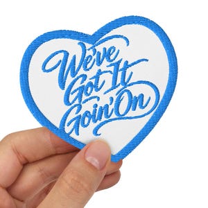 Könnte beinhalten: Ein herzförmiger Stickaufnäher mit weißem Hintergrund und einem leuchtend blauen Rand. Der Text "We've Got It Goin' On" ist in blauer Schreibschrift in die Mitte des Aufnähers gestickt und weist eine saubere, gestickte Stoffstruktur auf.