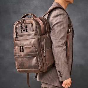 May include: A brown leather backpack with a large front pocket and a smaller pocket on the side. The backpack has a personalized monogram with the letter "M" and the name "Michael" on the front.
