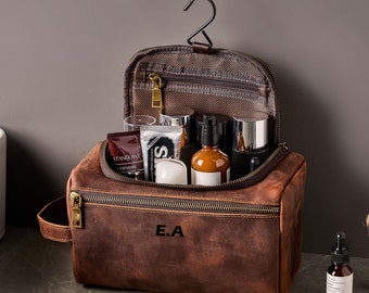 Puede incluir: Bolsa de aseo de cuero marrón con cierre de cremallera y asa. La bolsa está abierta y llena de diversos artículos de tocador, incluyendo botellas, tubos y frascos. La bolsa tiene las iniciales "E.A." grabadas en la parte delantera.
