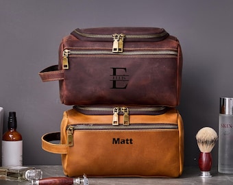 May include: Two brown leather toiletry bags with zipper closures. The top bag has the name "Ellis" and the initial "E" engraved on it. The bottom bag has the name "Matt" engraved on it.