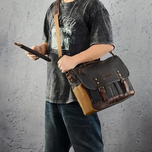Op de afbeelding: Een zwarte canvas messenger bag met bruine lederen details en riemen. De tas heeft een klepsluiting met gespen en een zandkleurig canvas zijpaneel. Hij wordt over de schouder gedragen met een bruine leren riem.