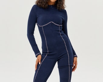 Könnte beinhalten: Ein marineblauer Jumpsuit mit kontrastierenden Nähten. Der Jumpsuit hat lange Ärmel und einen Stehkragen. Das Model trägt schwarze High-Top-Sneaker.