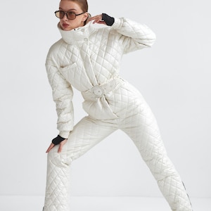 Puede incluir: Mono de nieve acolchado color crema con cuello alto, cinturón y botas negras. El conjunto de una pieza presenta un patrón de diamantes y una cremallera frontal. La modelo lleva gafas de sol y guantes negros.