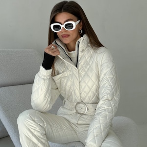 May include: A white quilted ski jumpsuit with a black belt and white oval sunglasses. The jumpsuit has a high collar and a zipper closure.