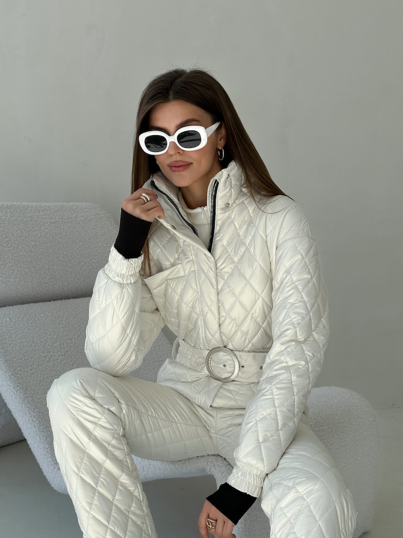 May include: A white quilted ski jumpsuit with a black belt and white oval sunglasses. The jumpsuit has a high collar and a zipper closure.