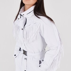 Puede incluir: Traje de esqu&iacute; blanco con ribete de flecos y cintur&oacute;n negro. El traje tiene cierre de cremallera y cuello alto.