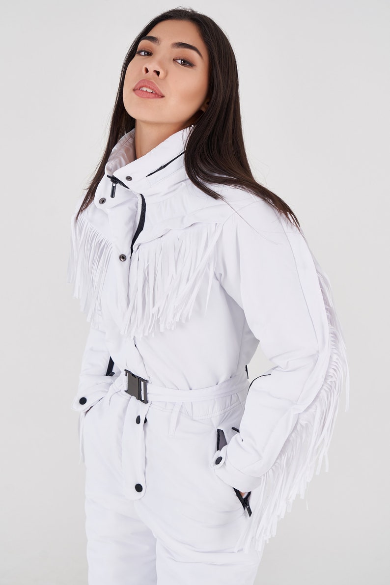 Puede incluir: Traje de esqu&iacute; blanco con ribete de flecos y cintur&oacute;n negro. El traje tiene cierre de cremallera y cuello alto.