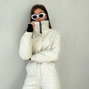 May include: A white quilted ski jumpsuit with a high collar and a zipper closure. The jumpsuit has a diamond pattern and a black belt. The model is wearing white sunglasses.