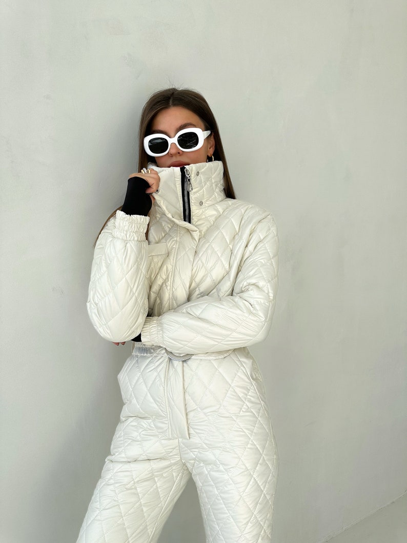 May include: A white quilted ski jumpsuit with a high collar and a zipper closure. The jumpsuit has a diamond pattern and a black belt. The model is wearing white sunglasses.