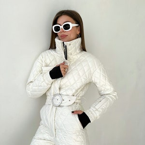 May include: A white quilted ski jumpsuit with a black belt and white buckle. The jumpsuit has a high collar and a zipper closure. The model is wearing white sunglasses.