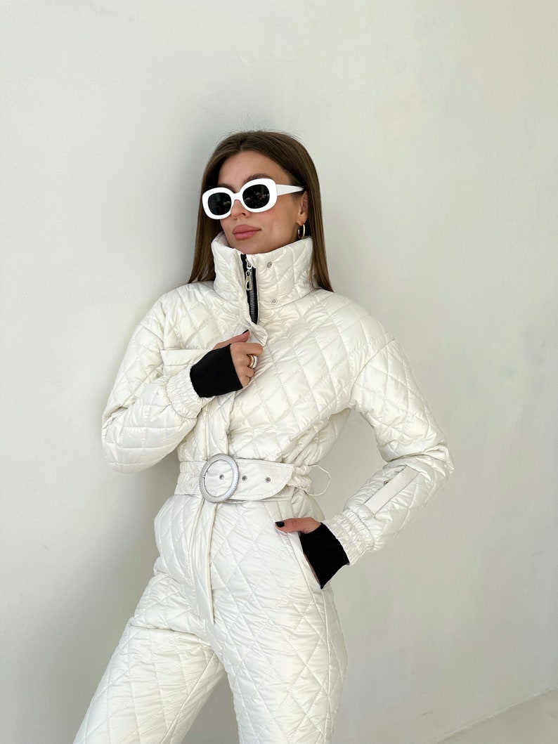 May include: A white quilted ski jumpsuit with a black belt and white buckle. The jumpsuit has a high collar and a zipper closure. The model is wearing white sunglasses.
