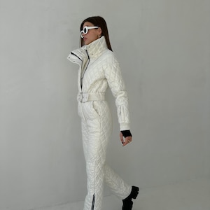 May include: A white quilted ski jumpsuit with a black zipper and a white belt. The jumpsuit has a high collar and a zipper closure. The model is wearing white sunglasses.