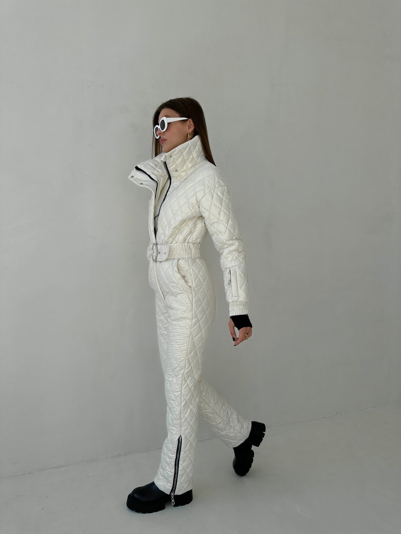 May include: A white quilted ski jumpsuit with a black zipper and a white belt. The jumpsuit has a high collar and a zipper closure. The model is wearing white sunglasses.