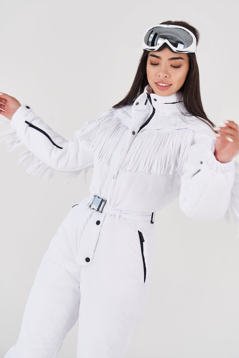 Puede incluir: Un mono de esqu&iacute; blanco con un detalle de flecos en el pecho y las mangas. El mono tiene un cintur&oacute;n negro y una cremallera. La modelo lleva unas gafas de esqu&iacute; blancas.