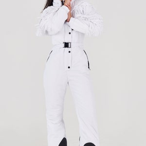 Puede incluir: Un mono de esqu&iacute; blanco con cintur&oacute;n negro y detalles negros en las piernas. El mono tiene un detalle de flecos en las mangas.