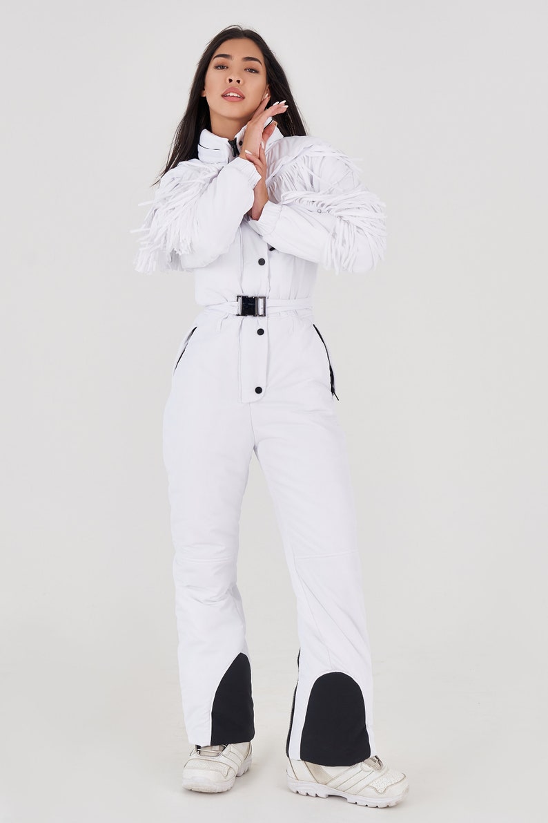 Puede incluir: Un mono de esqu&iacute; blanco con cintur&oacute;n negro y detalles negros en las piernas. El mono tiene un detalle de flecos en las mangas.