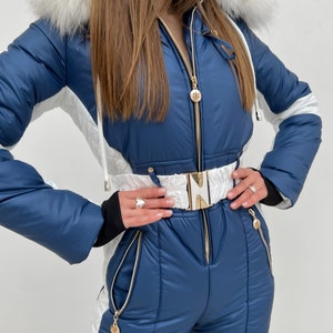 K&ouml;nnte beinhalten: Ein blauer und wei&szlig;er Ski-Overall mit einem wei&szlig;en Pelzbesatz an der Kapuze. Der Overall hat einen goldenen Rei&szlig;verschluss und eine goldene G&uuml;rtelschnalle.