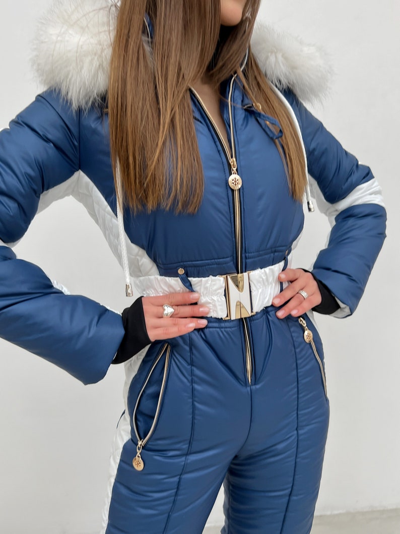 K&ouml;nnte beinhalten: Ein blauer und wei&szlig;er Ski-Overall mit einem wei&szlig;en Pelzbesatz an der Kapuze. Der Overall hat einen goldenen Rei&szlig;verschluss und eine goldene G&uuml;rtelschnalle.