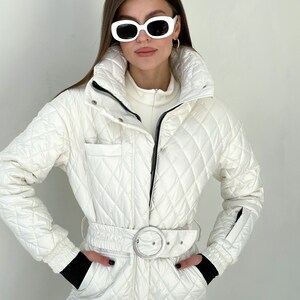 May include: A white quilted ski jumpsuit with a black belt and a silver buckle. The jumpsuit has a zipper closure and a high collar.