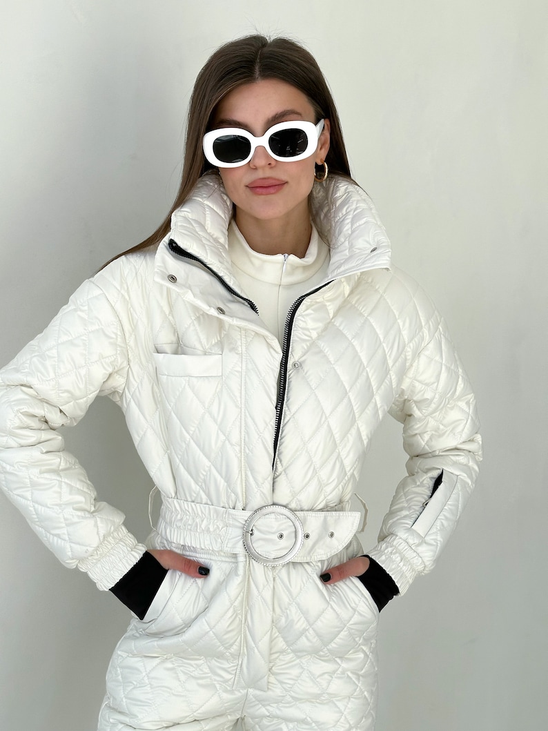 May include: A white quilted ski jumpsuit with a black belt and a silver buckle. The jumpsuit has a zipper closure and a high collar.